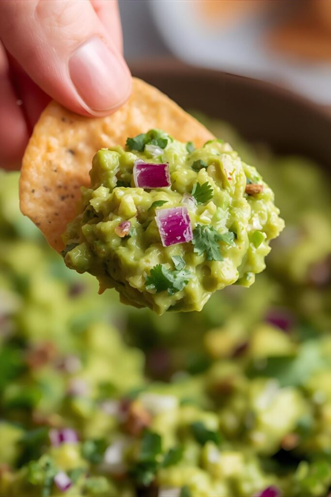 A very close-up, appetizing shot of a scoop of chunky Chipotle guacamole on a crisp tortilla chip, showing the texture of the avocado, cilantro, and red onion, with a blurred background of a full bowl of guac. `