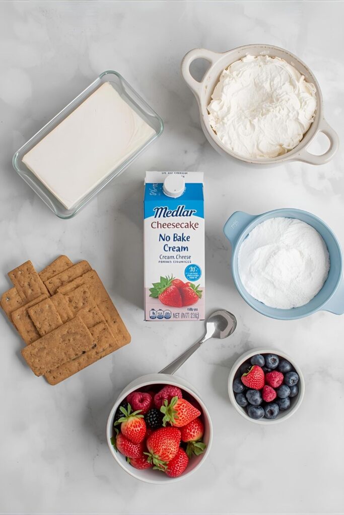 A flat lay of no-bake cheesecake ingredients including cream cheese, graham crackers, heavy cream, and berries on a bright countertop.