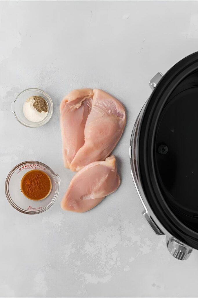 A flat-lay image of the ingredients for slow cooker chicken, including raw chicken breasts, spices, and broth, arranged next to a Crock-Pot.