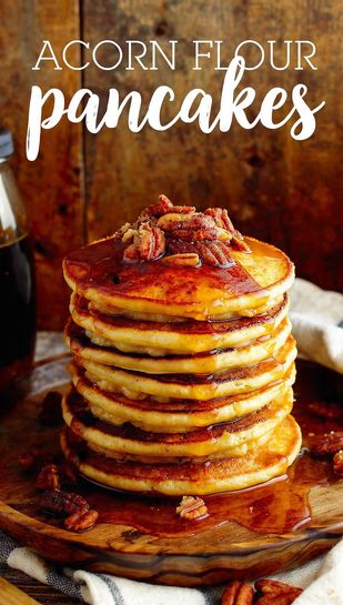 A tall stack of golden-brown acorn flour pancakes, drizzled with maple syrup and topped with pecans, set on a rustic wooden table. Text overlay reads: ACORN FLOUR PANCAKES