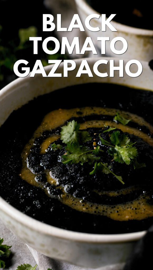 A close-up shot of a bowl of dark, rich Black Tomato Gazpacho, garnished with finely diced green bell pepper, red onion, and fresh basil, with a drizzle of olive oil. The text overlay reads: "BLACK TOMATO GAZPACHO"