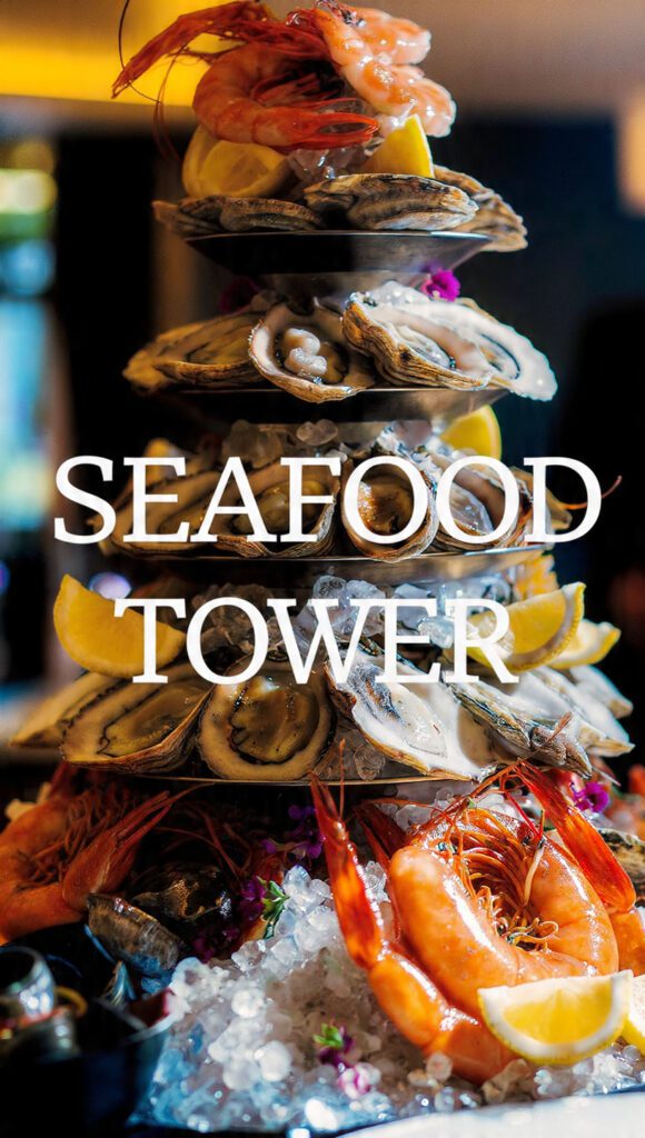 Close-up of a tiered Seafood Tower with shrimp, oysters, and crab on crushed ice. Text overlay reads "SEAFOOD TOWER".