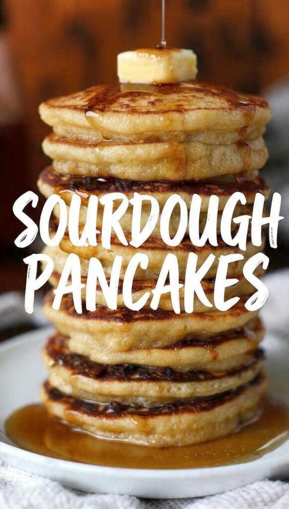 A tall, perfect stack of thick, golden-brown Sourdough Pancakes, topped with a pat of butter and thick maple syrup dripping down the sides. The texture shows small, delicate air bubbles. The text overlay reads: "SOURDOUGH PANCAKES".