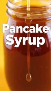 A glass jar of golden brown pancake syrup being poured over a stack of pancakes with text overlay "Pancake Syrup".
